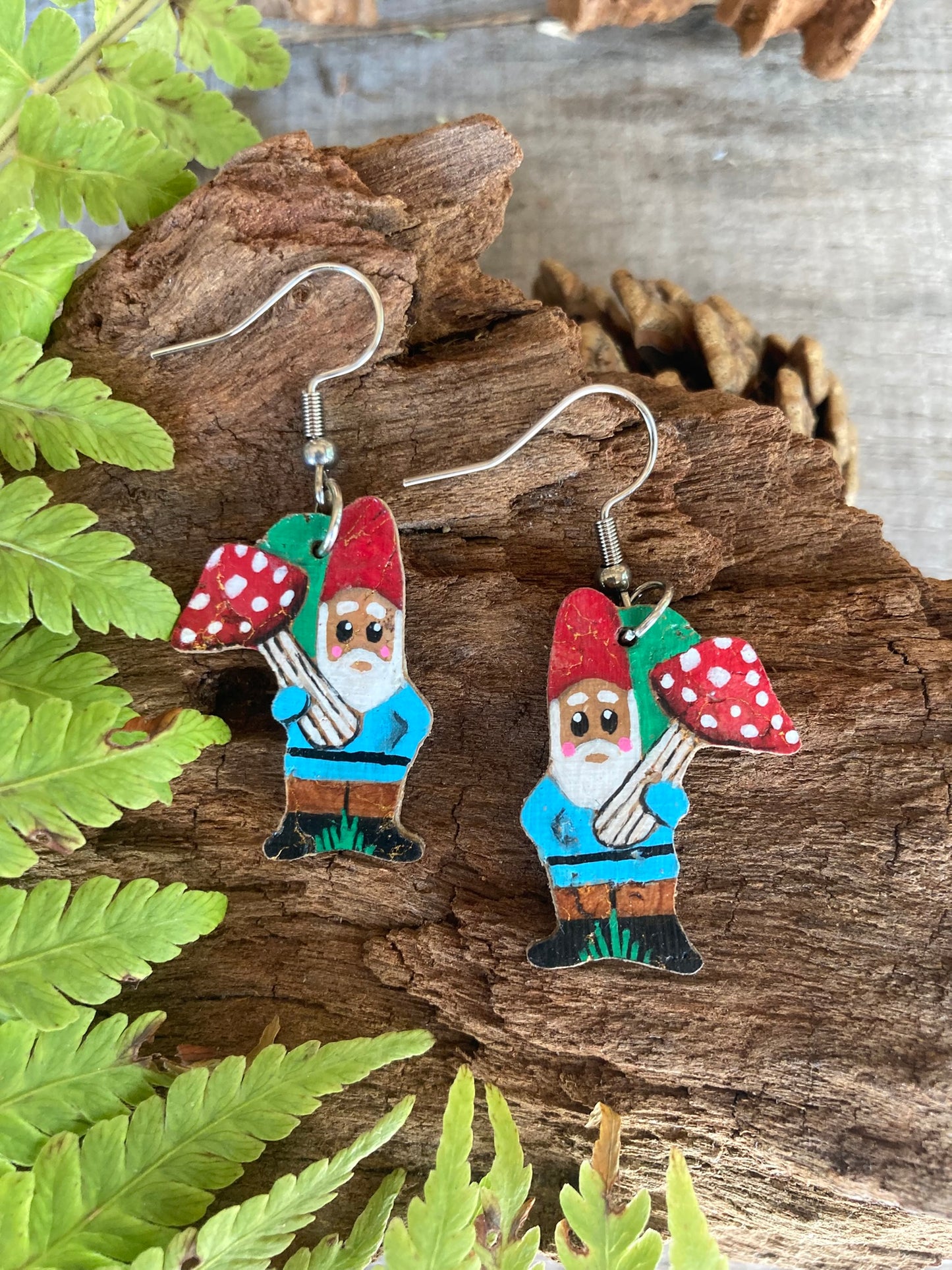 Gnome earrings with red hats and blue outfits on a wooden surface with fern leaves.