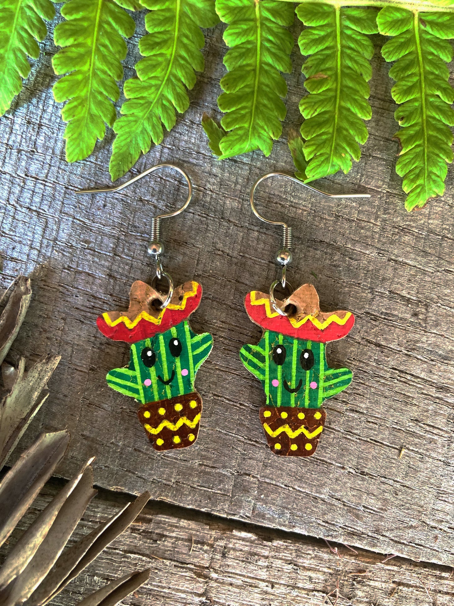 Cactus-shaped earrings with sombreros on a wooden surface with green leaves.