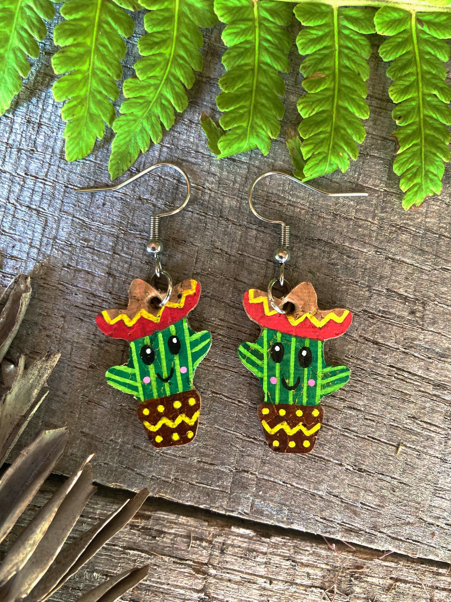 Cactus-shaped earrings with sombreros on a wooden surface with green leaves.