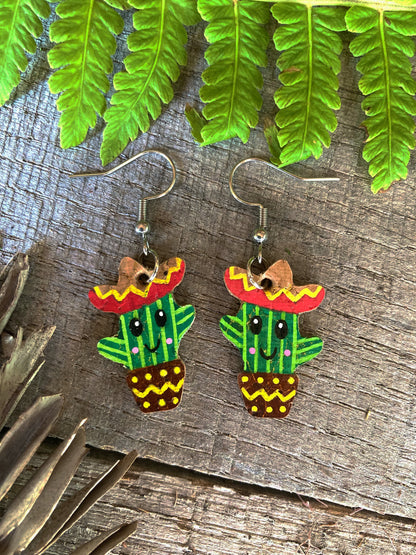 Cactus-shaped earrings with sombreros on a wooden surface with green leaves.