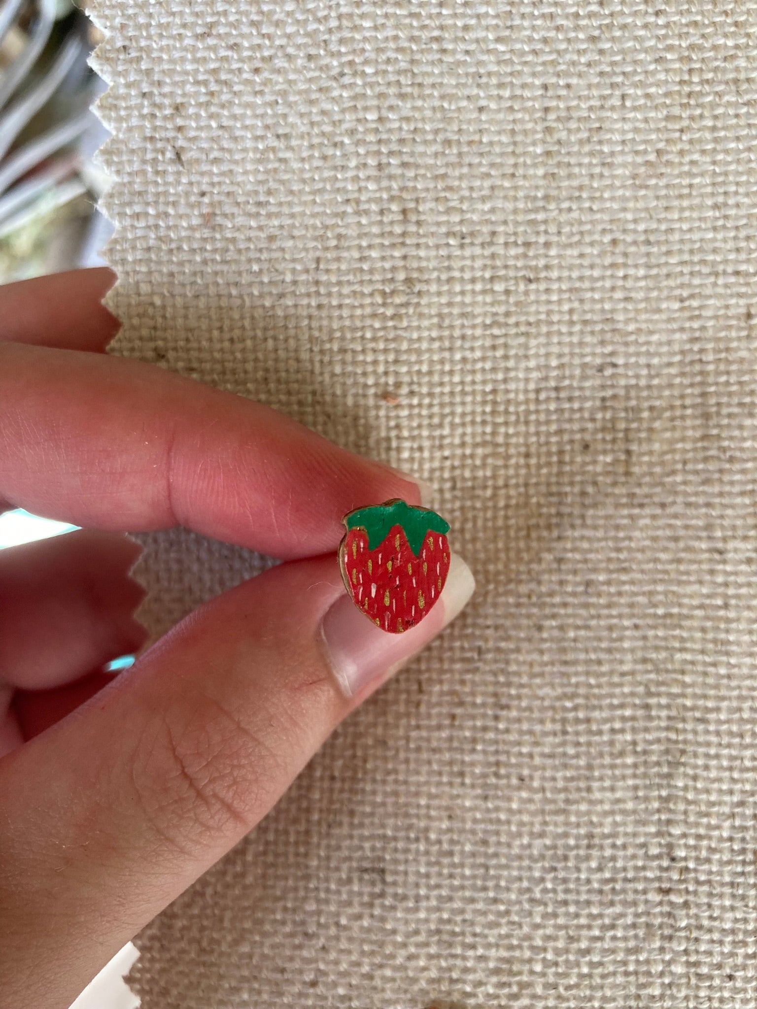 Small strawberry-shaped stud earrings held between fingers on a textured fabric background