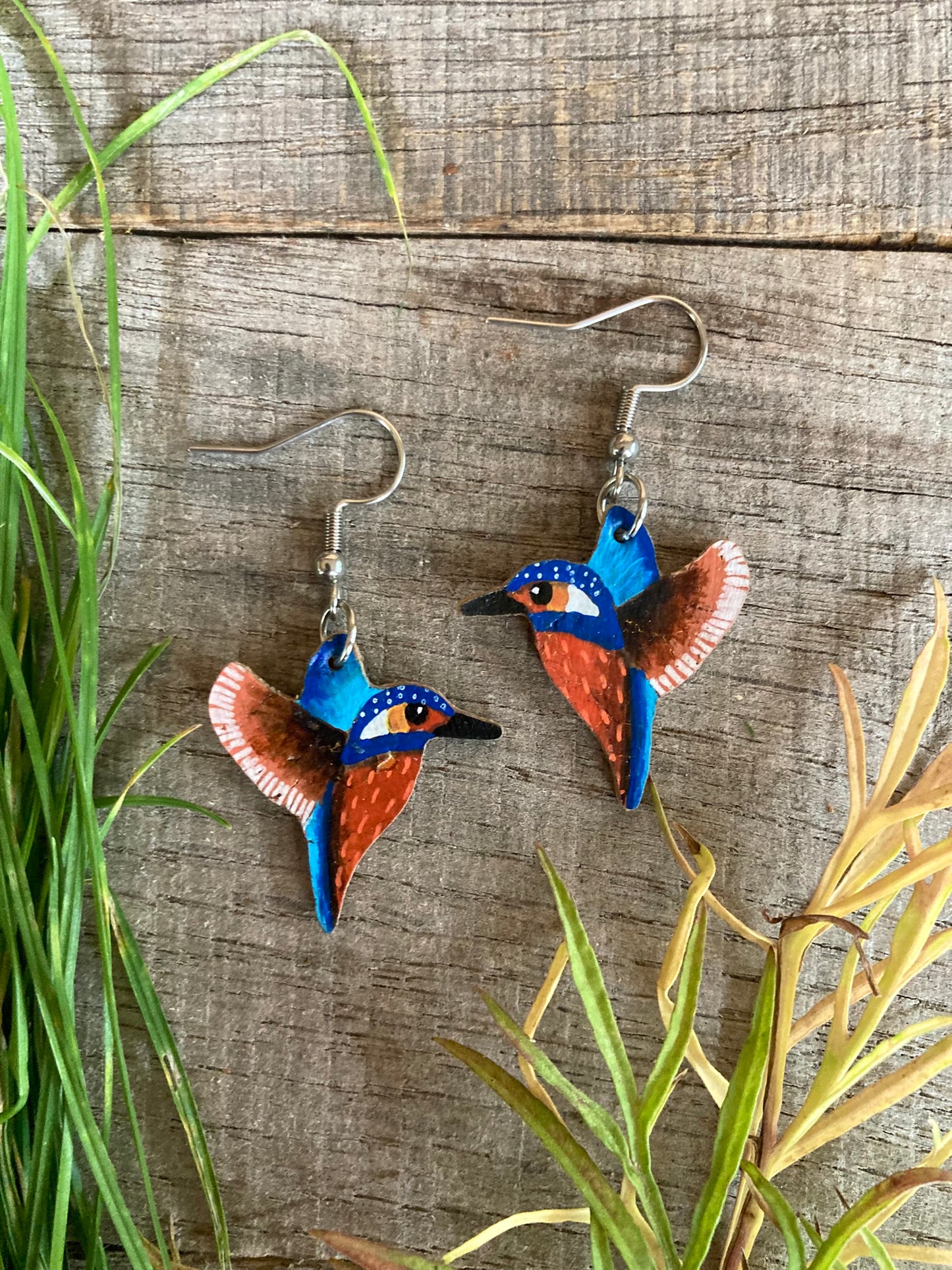 Blue and orange kingfisher earrings on a wooden surface with grass and leaves.