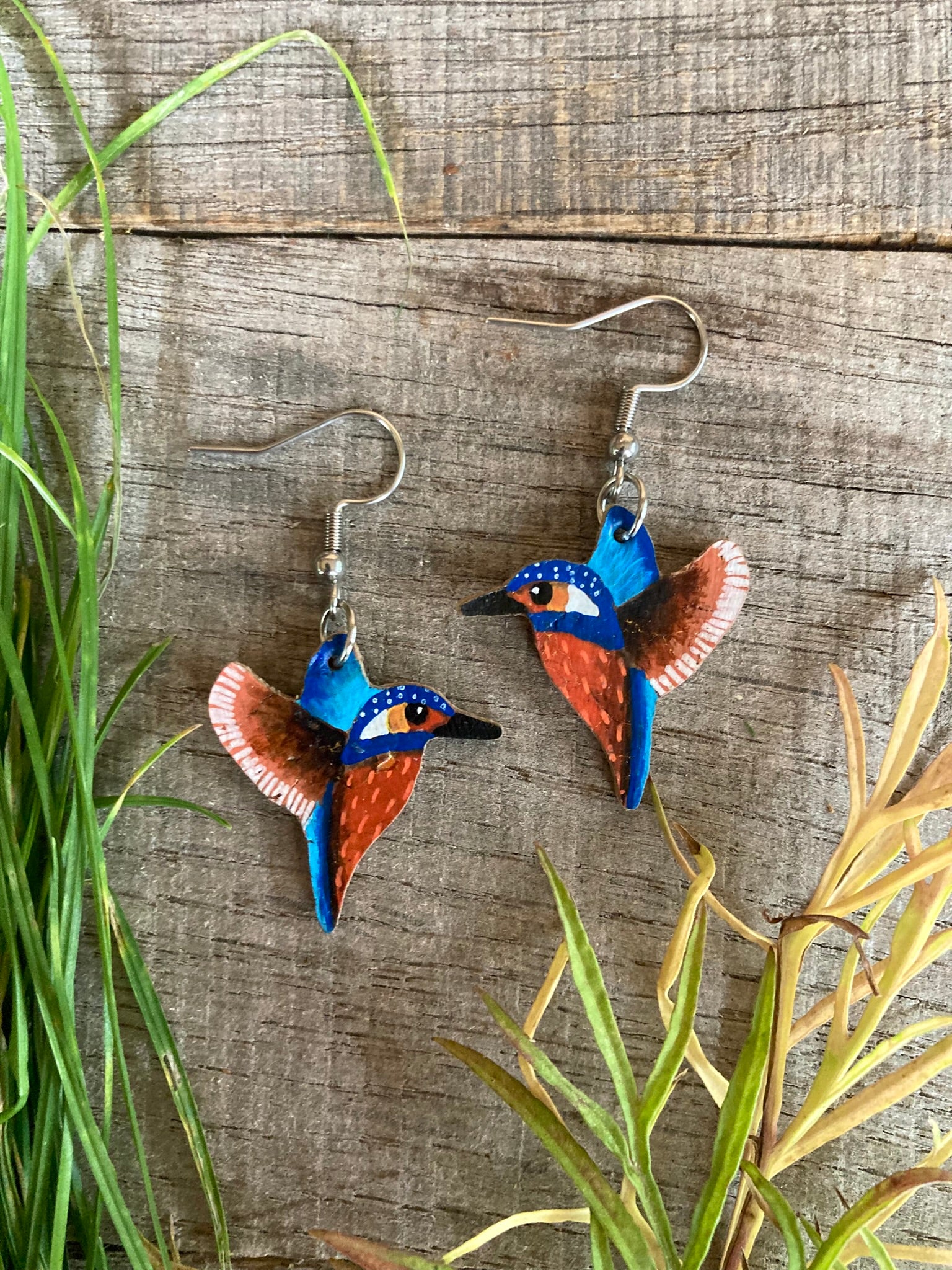 Blue and orange kingfisher earrings on a wooden surface with grass and leaves.