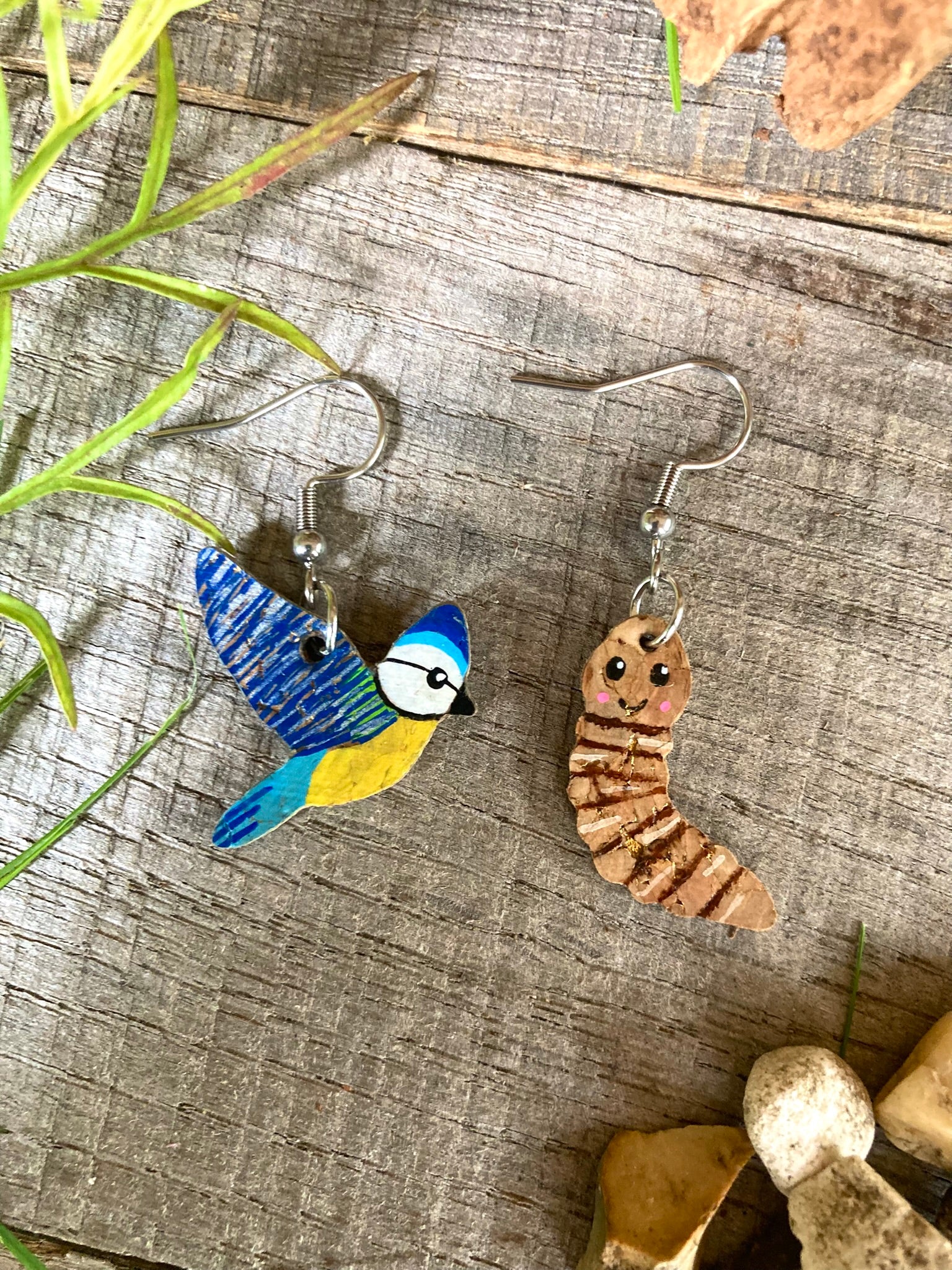 Bird and worm-shaped earrings on a wooden surface with plants.