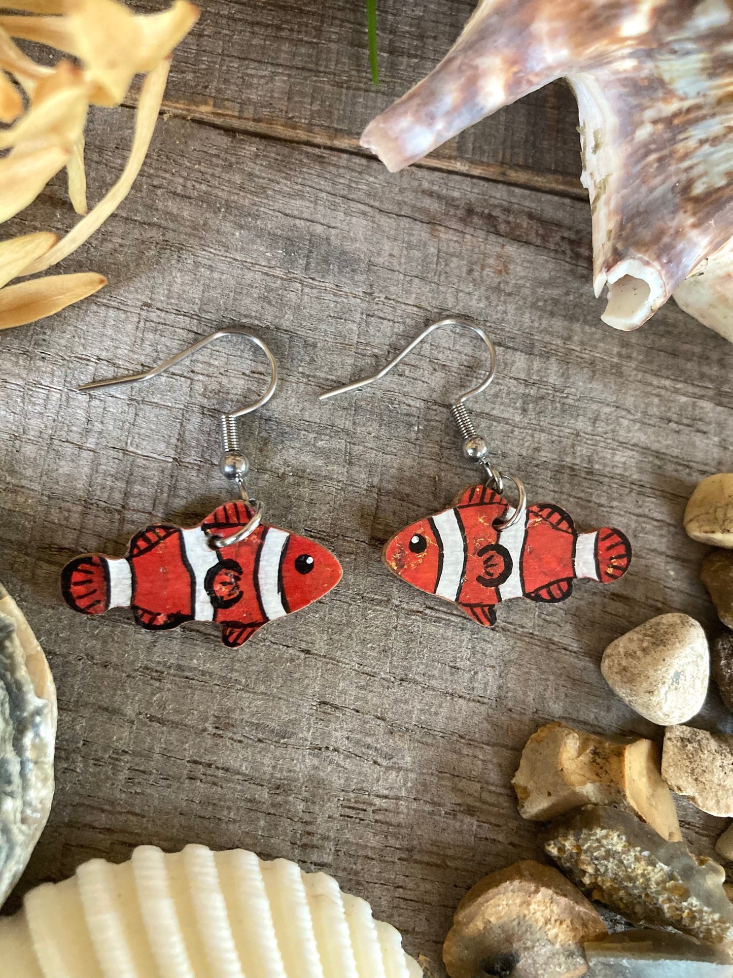 Pair of clownfish earrings on a wooden surface with shells and stones.