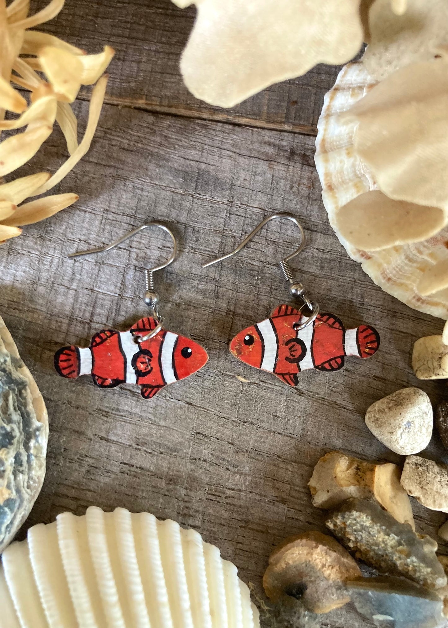 Nemo fish earrings on a wooden surface with seashells and stones.