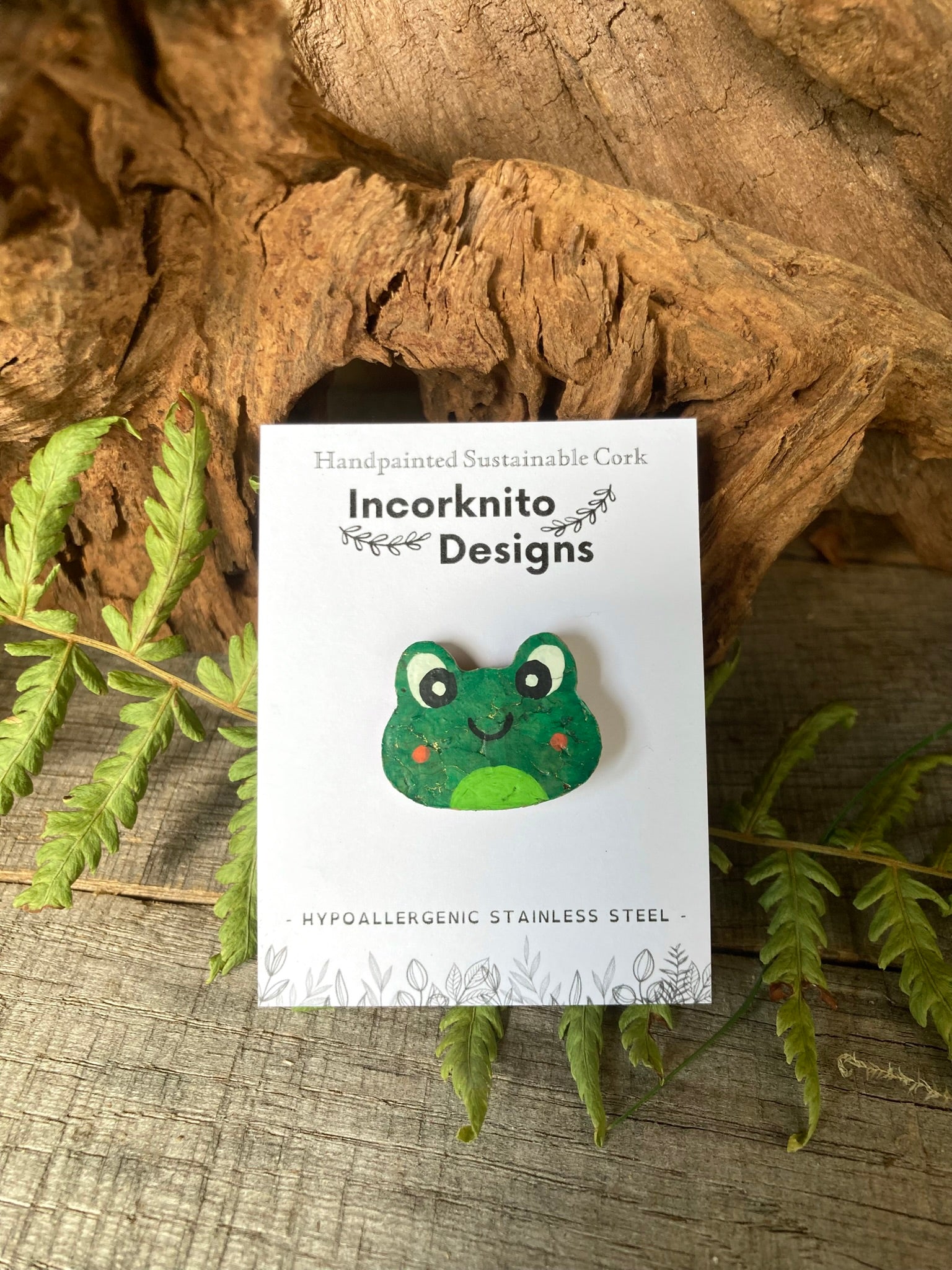 Green frog-shaped pin badge on a white card with 'Incorkinito Designs' branding, placed on a natural wood and leaf background.