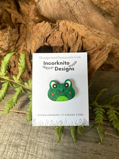 Green frog-shaped pin badge on a white card with 'Incorkinito Designs' branding, placed on a natural wood and leaf background.