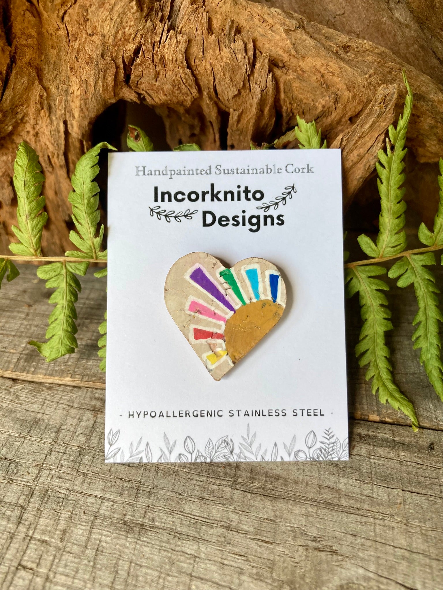 Heart-shaped cork pin with colourful design on a white card with 'Incorknito Designs' branding, placed on a wooden surface with green leaves.