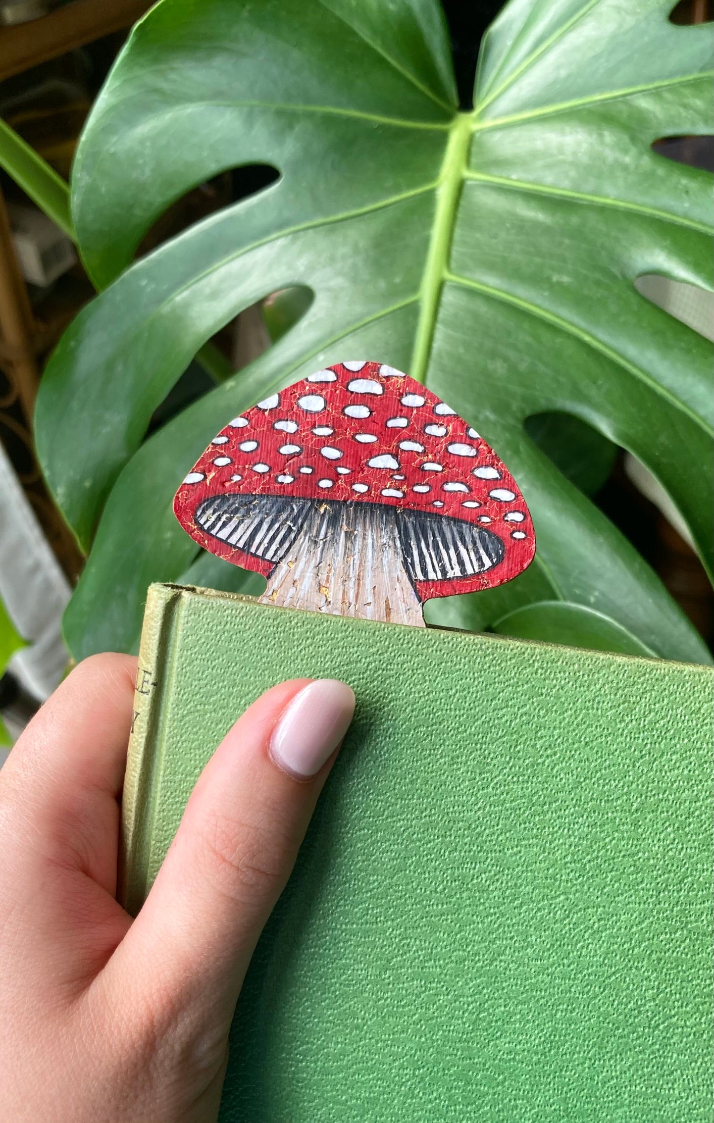 Hand holding a green book with a red and white mushroom-shaped bookmark against a leafy background