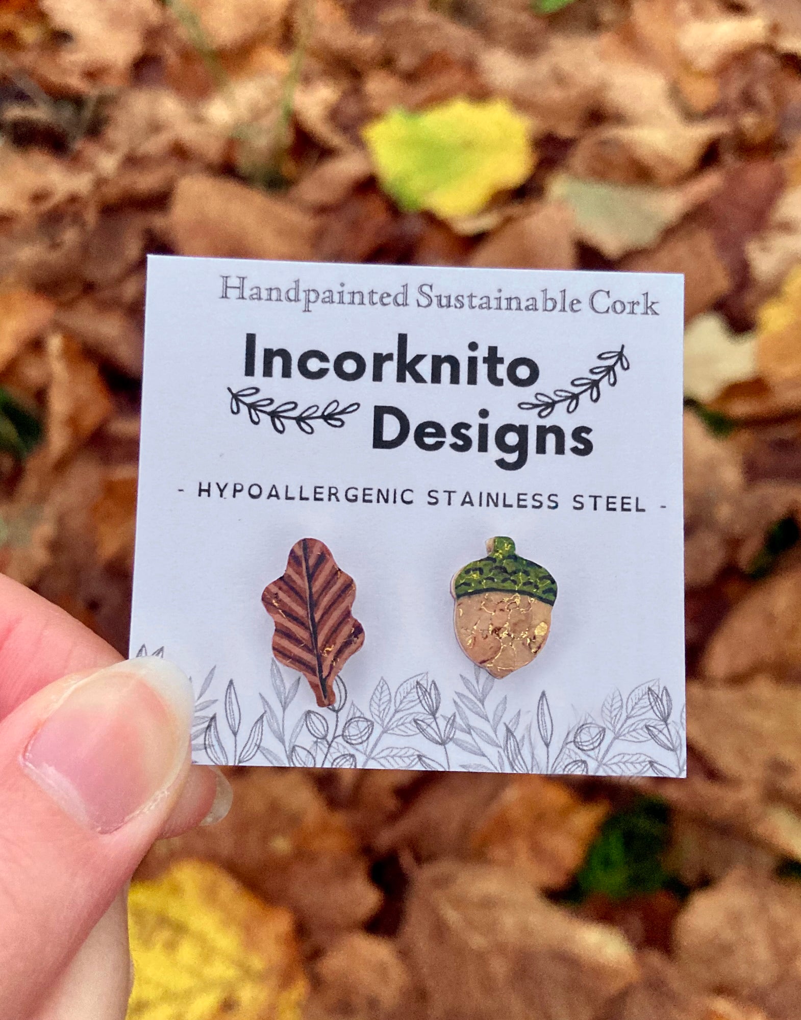 Handpainted acorn and oak leaf mismatched cork stud earrings on an Incorknito Designs card held against a carpet of autumn leaves