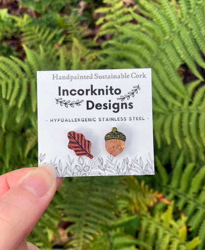 Handpainted acorn and oak leaf mismatched cork stud earrings on an Incorknito Designs card held against green fern fronds