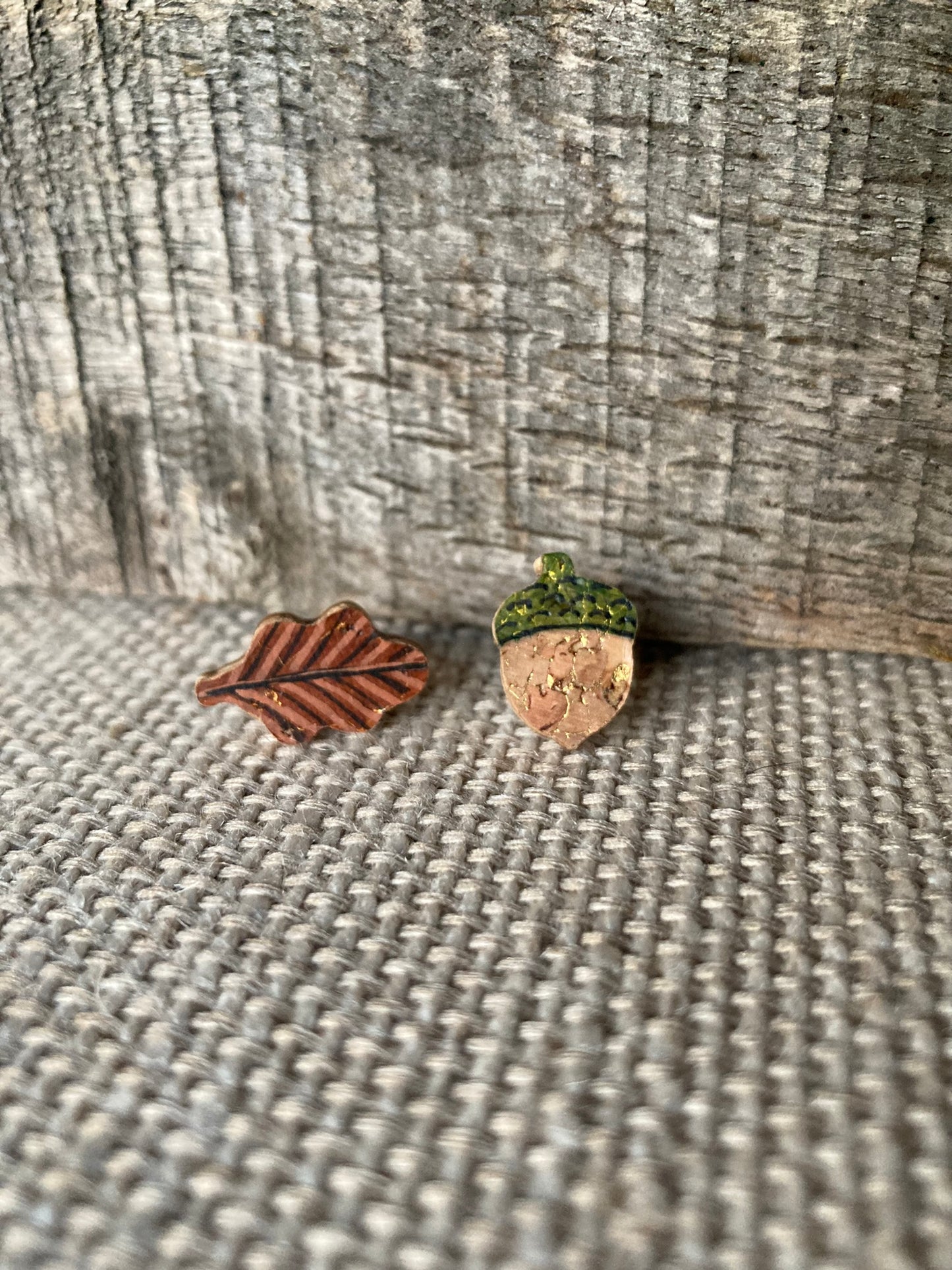 Acorn and oak leaf cork stud earrings on a linen surface against weathered wood, showing fine vein detail on the leaf and gold fleck acorn body
