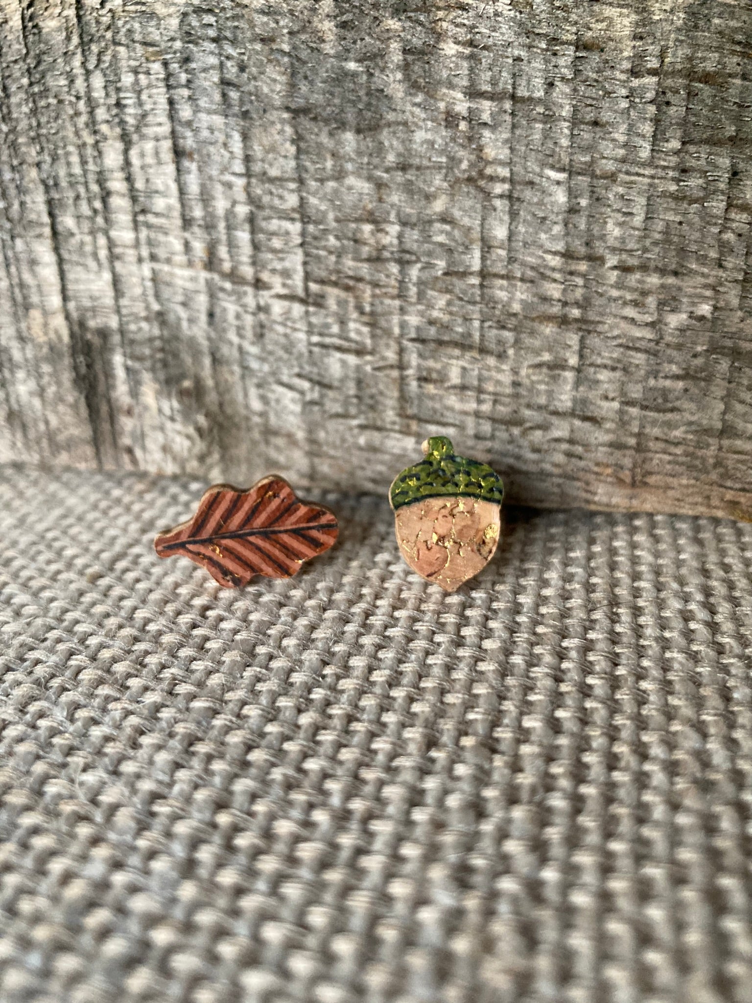 Acorn and oak leaf cork stud earrings on a linen surface against weathered wood, showing fine vein detail on the leaf and gold fleck acorn body