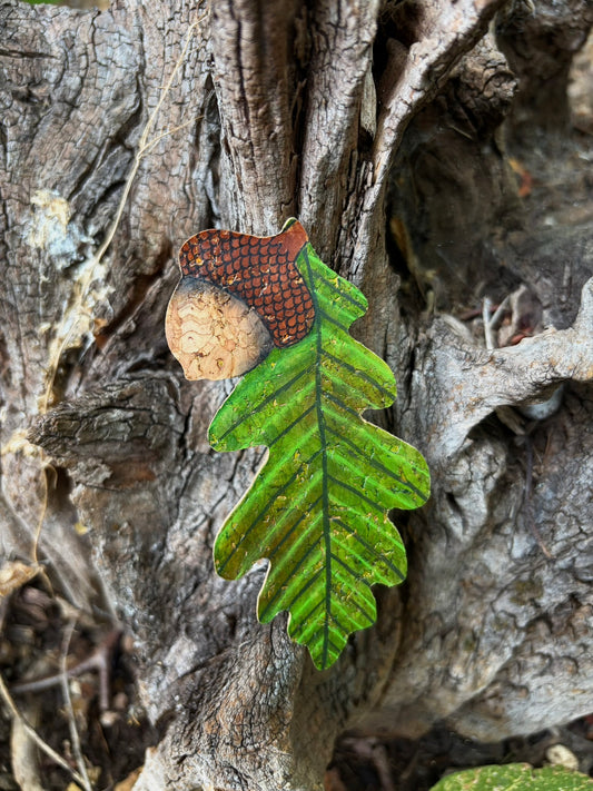 Handpainted acorn and oak leaf cork bookmark against oak tree bark — rich green lobed leaf with fine vein detail and gold fleck shimmer, natural cork acorn with dark brown crosshatch cap