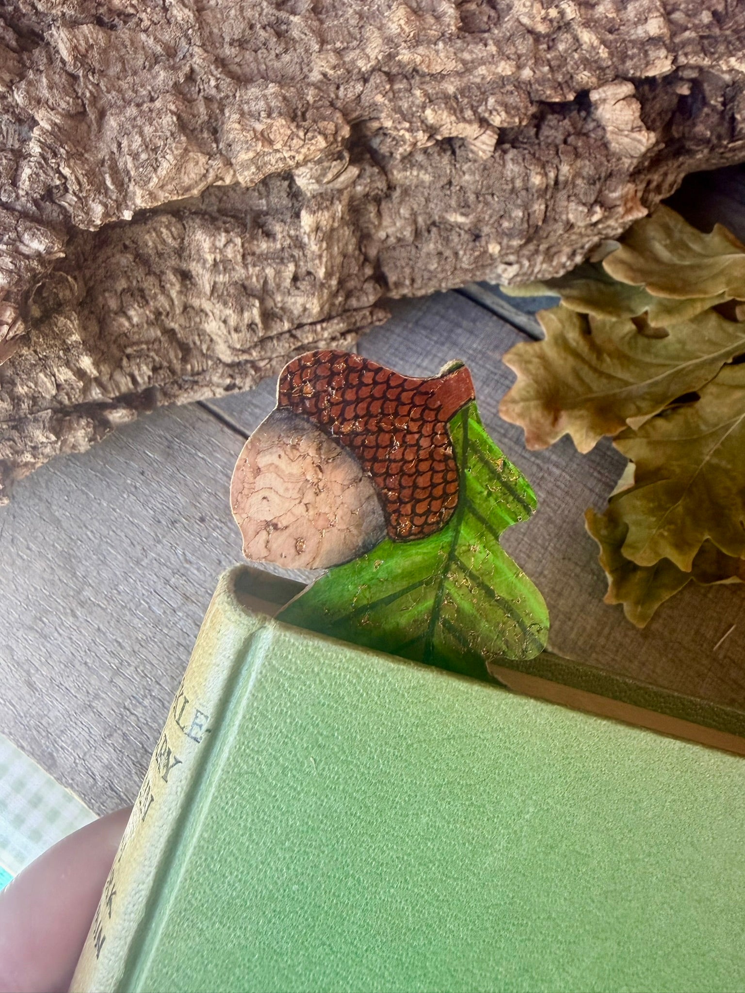 Acorn and oak leaf cork bookmark peeking out of a green hardback book with cork bark and dried oak leaves — handpainted sustainable cork bookmark
