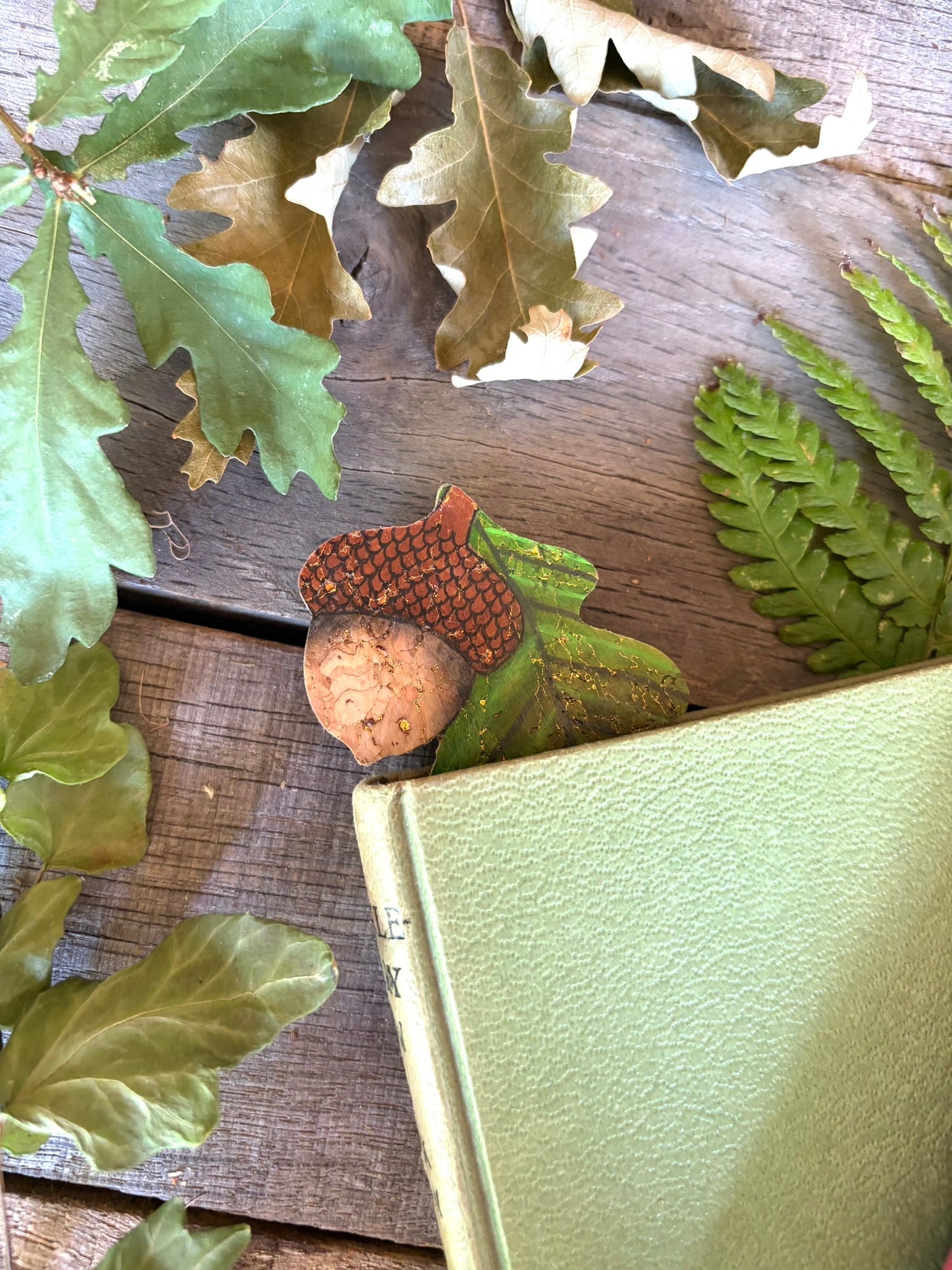 Acorn and oak leaf cork bookmark peeking out of a green hardback book with oak leaves and fern fronds — handpainted sustainable cork bookmark
