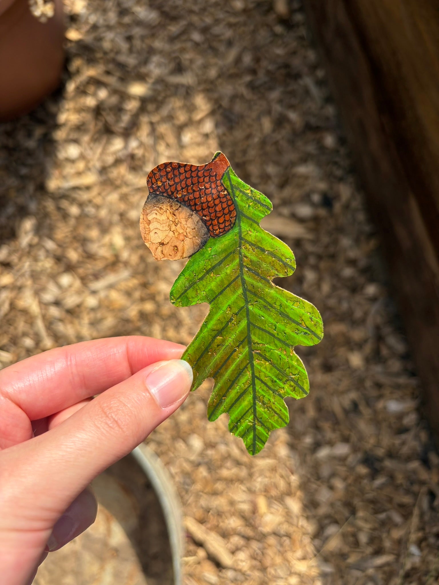 Handpainted acorn and oak leaf cork bookmark held up against a bark mulch backdrop — green lobed oak leaf with natural cork acorn body