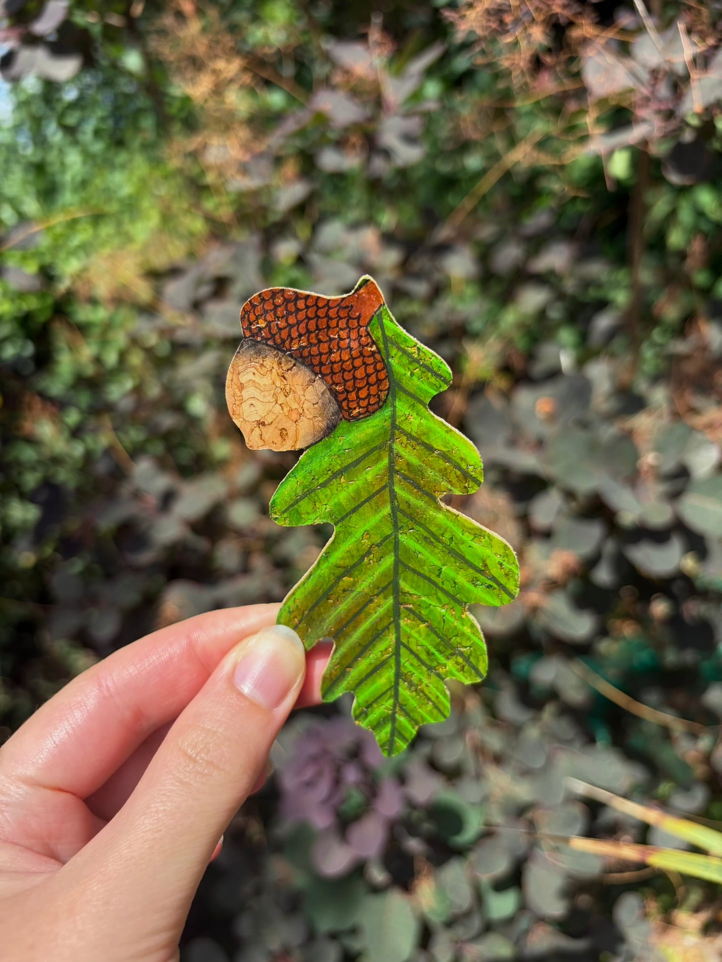 Handpainted acorn and oak leaf cork bookmark held against green ivy — rich green oak leaf with gold fleck shimmer and natural cork acorn