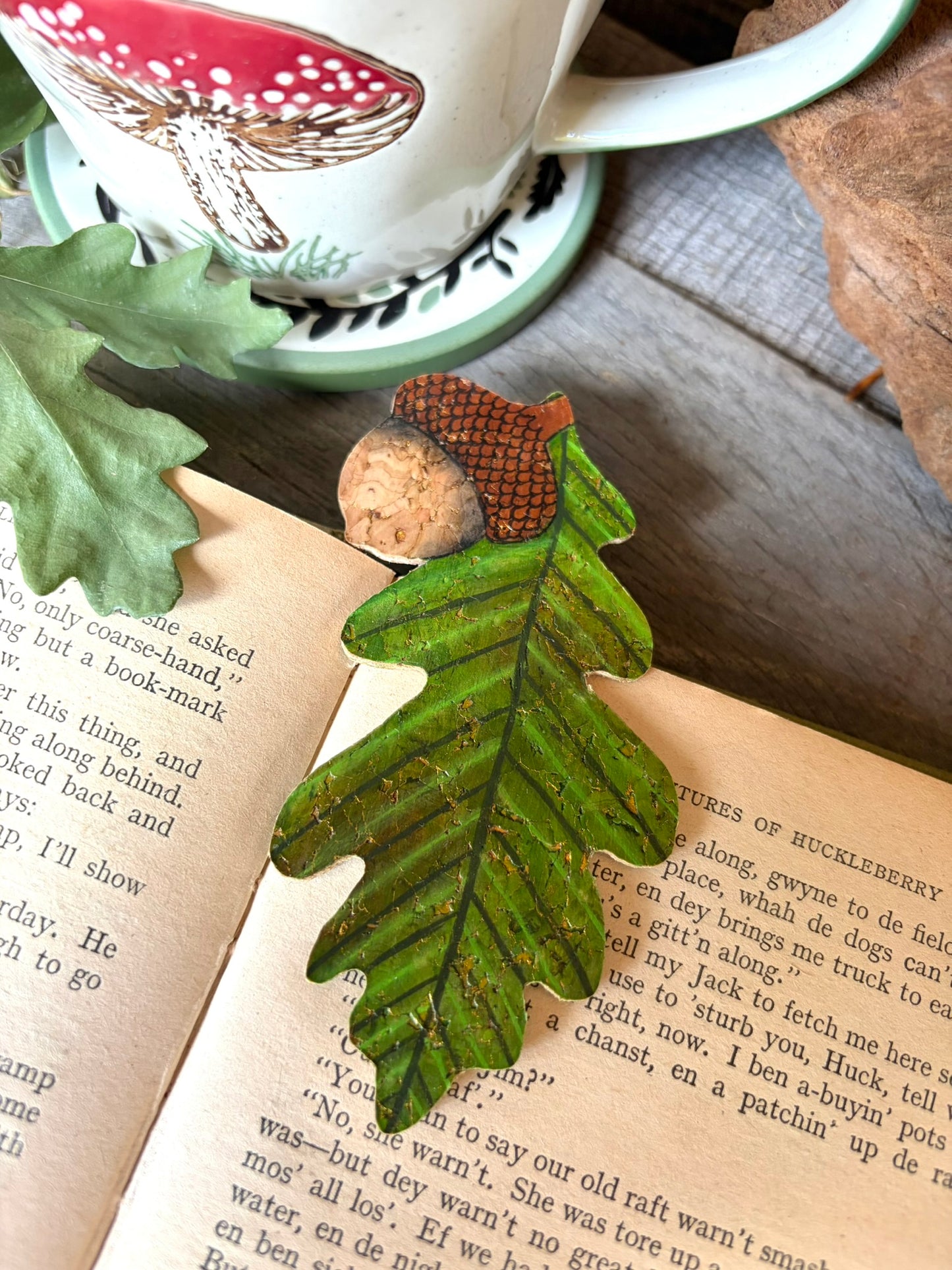 Acorn and oak leaf cork bookmark propped on an open book spine with a toadstool mug in the background — showing double-sided design, handpainted sustainable cork
