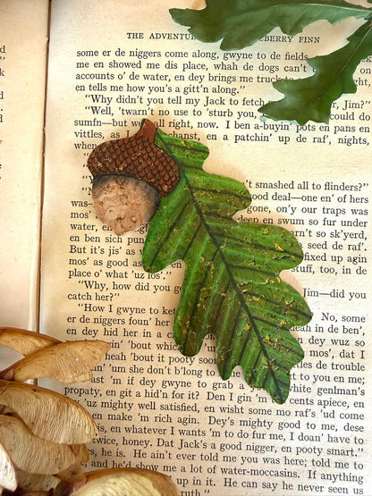 Acorn and oak leaf cork bookmark laid on open vintage book pages — handpainted green oak leaf with gold fleck shimmer and cork acorn