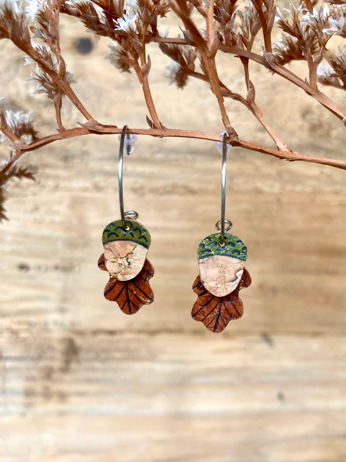 Acorn and oak leaf cork mini hoop earrings hanging from a branch with a natural wood plank background