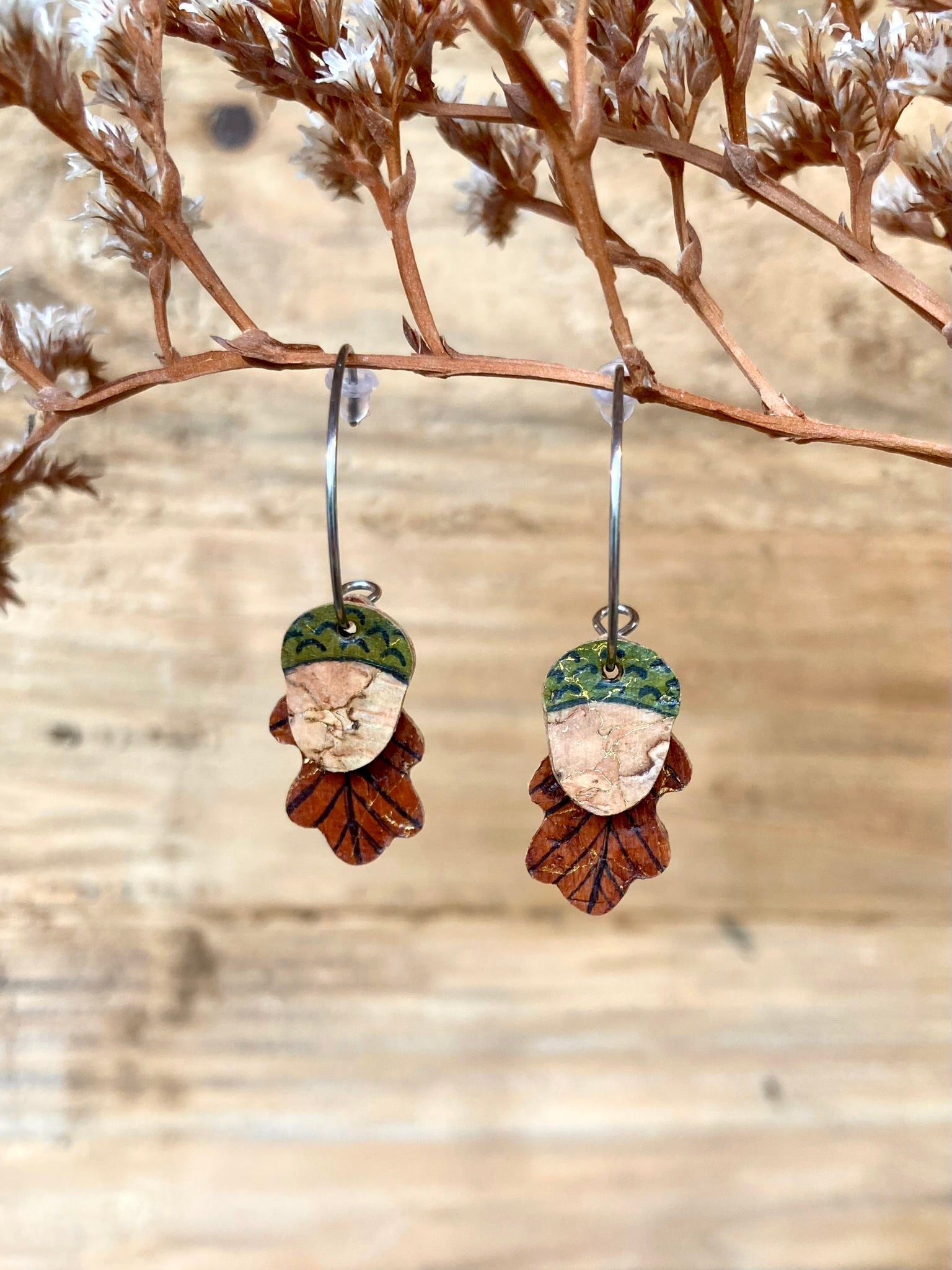 Acorn and oak leaf cork mini hoop earrings hanging from a branch with a natural wood plank background