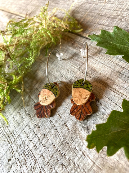 Handpainted cork acorn and oak leaf mini hoop earrings on a weathered wooden surface with fresh oak leaves and moss