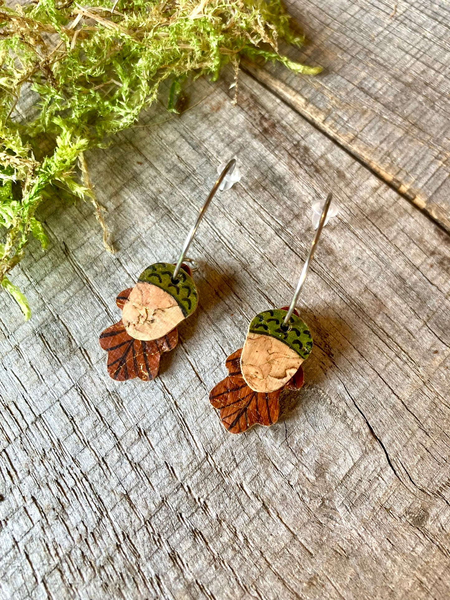 Close-up of handpainted acorn and oak leaf cork earrings on silver open-back mini hoops, laid on a wooden surface with moss