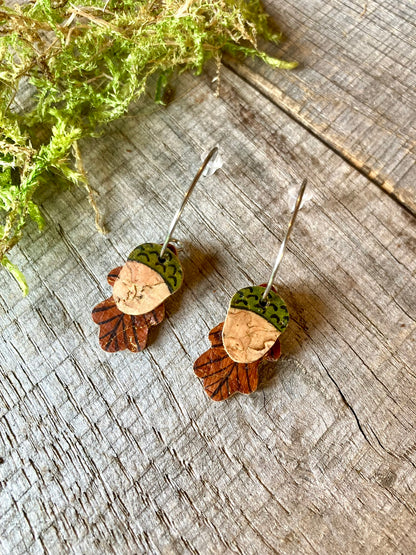Close-up of handpainted acorn and oak leaf cork earrings on silver open-back mini hoops, laid on a wooden surface with moss