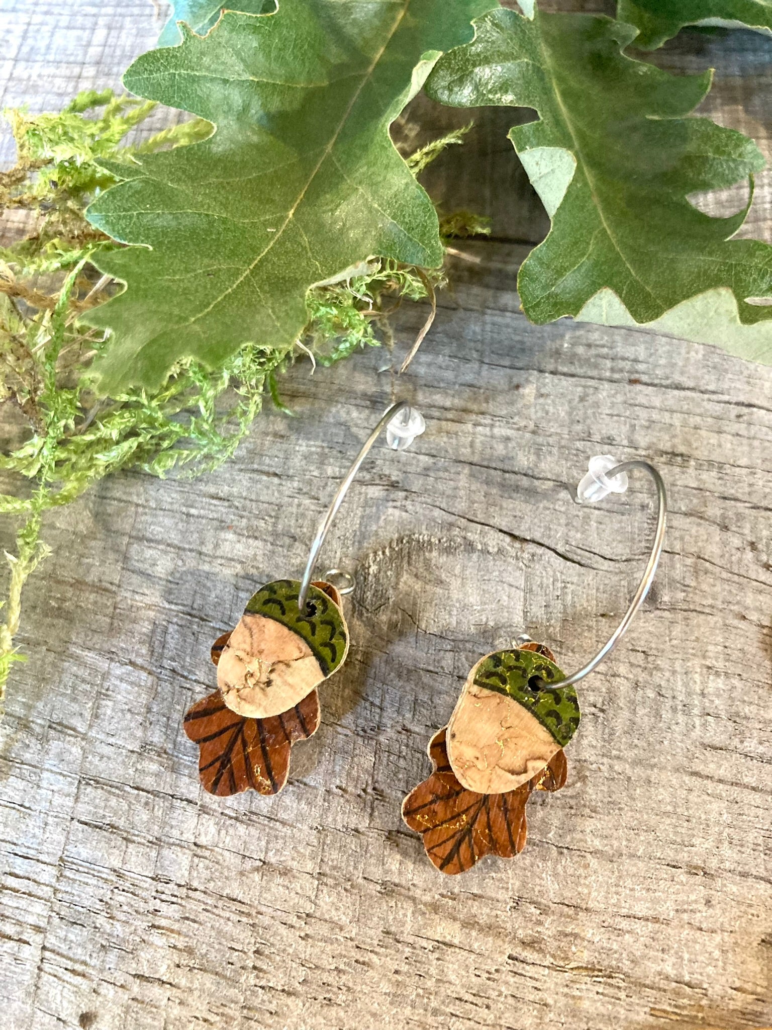 Handpainted cork acorn earrings on silver open-back mini hoops, displayed on a wooden surface with fresh green oak leaves