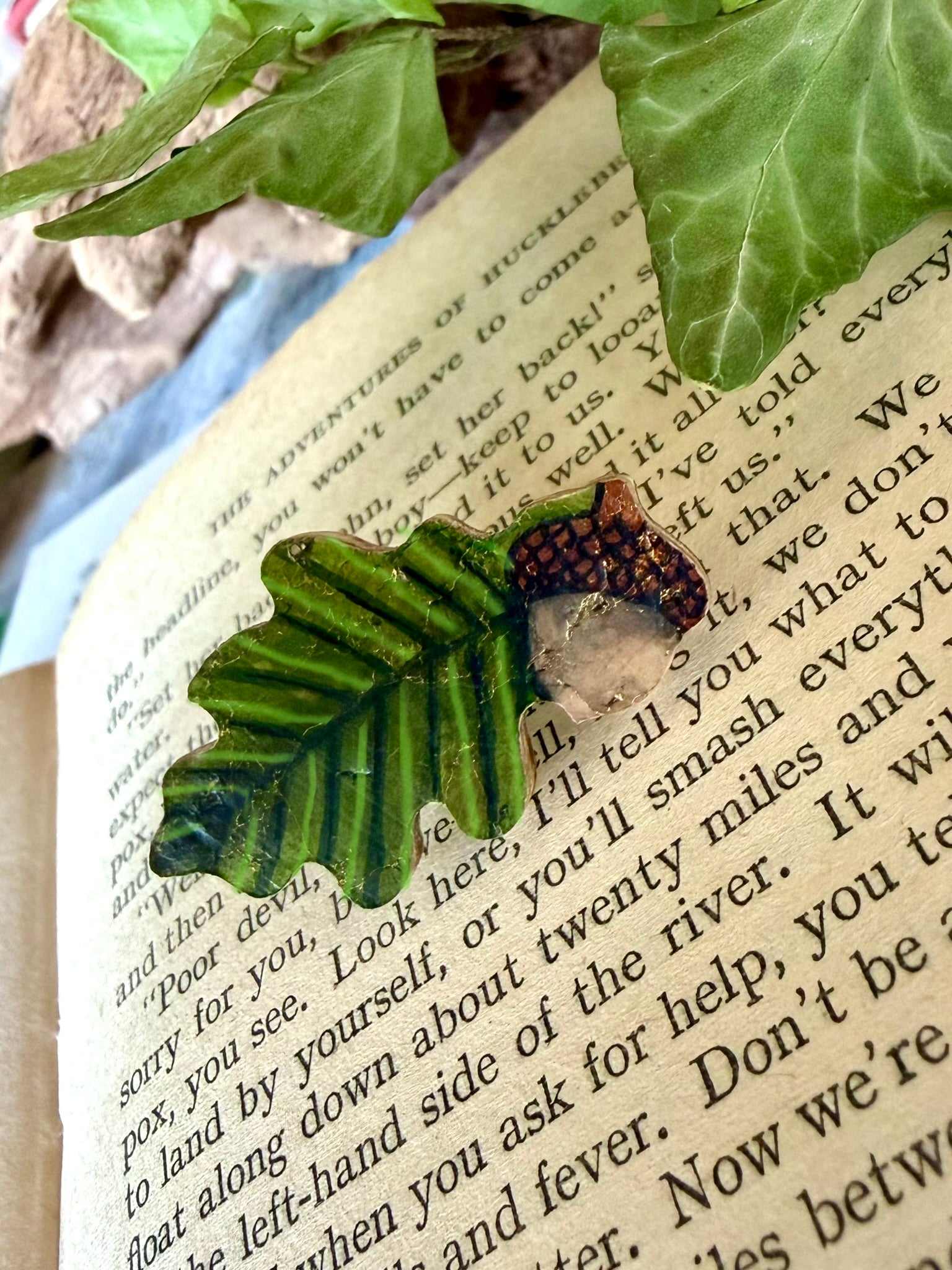 Handpainted acorn and oak leaf cork pin badge resting on open book pages with ivy leaves — moss green leaf with gold flecks and earthy brown acorn cap