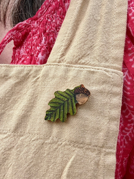 Handpainted acorn and oak leaf cork pin badge pinned to a linen dungaree pocket — moss green oak leaf with gold cork flecks and natural beige acorn, handmade in Suffolk UK
