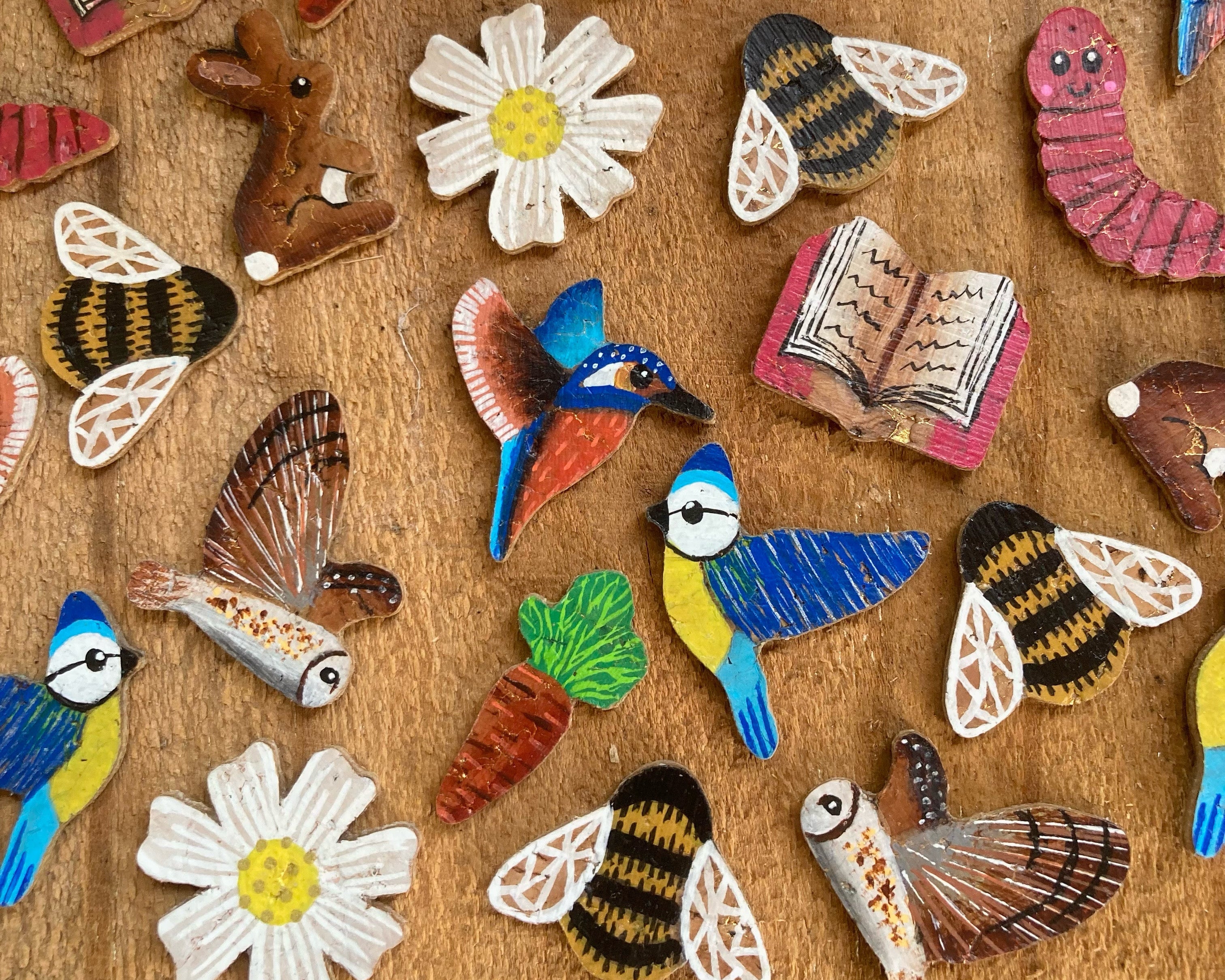 A collection of colourful hand-painted cork earrings pieces mid production, including kingfishers, bluetits, bees, flowers, books, and carrots, arranged on a textured brown wooden surface.