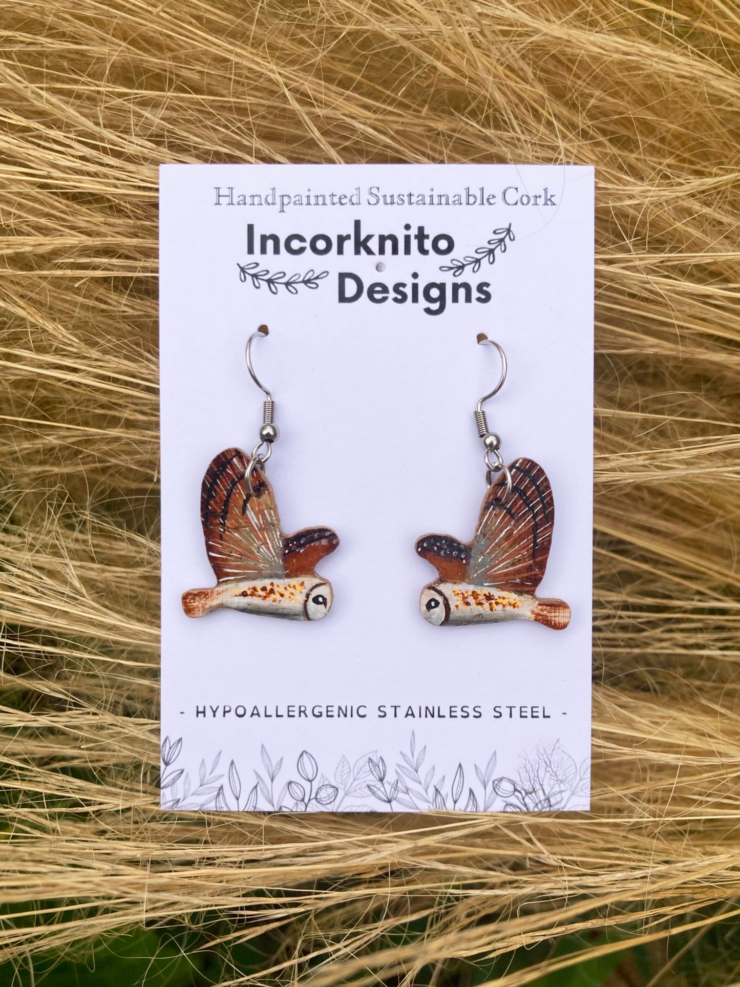 Barn owl cork earrings on silver hooks on Incorknito Designs card against pale golden grass — handpainted British wildlife cork earrings
