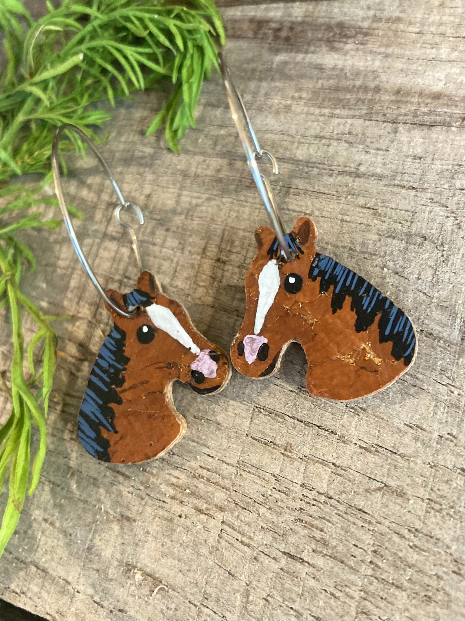 Bay brown horse cork earrings on large silver hoops, flat lay on weathered wood with greenery — eco-friendly handpainted horse jewellery Suffolk UK