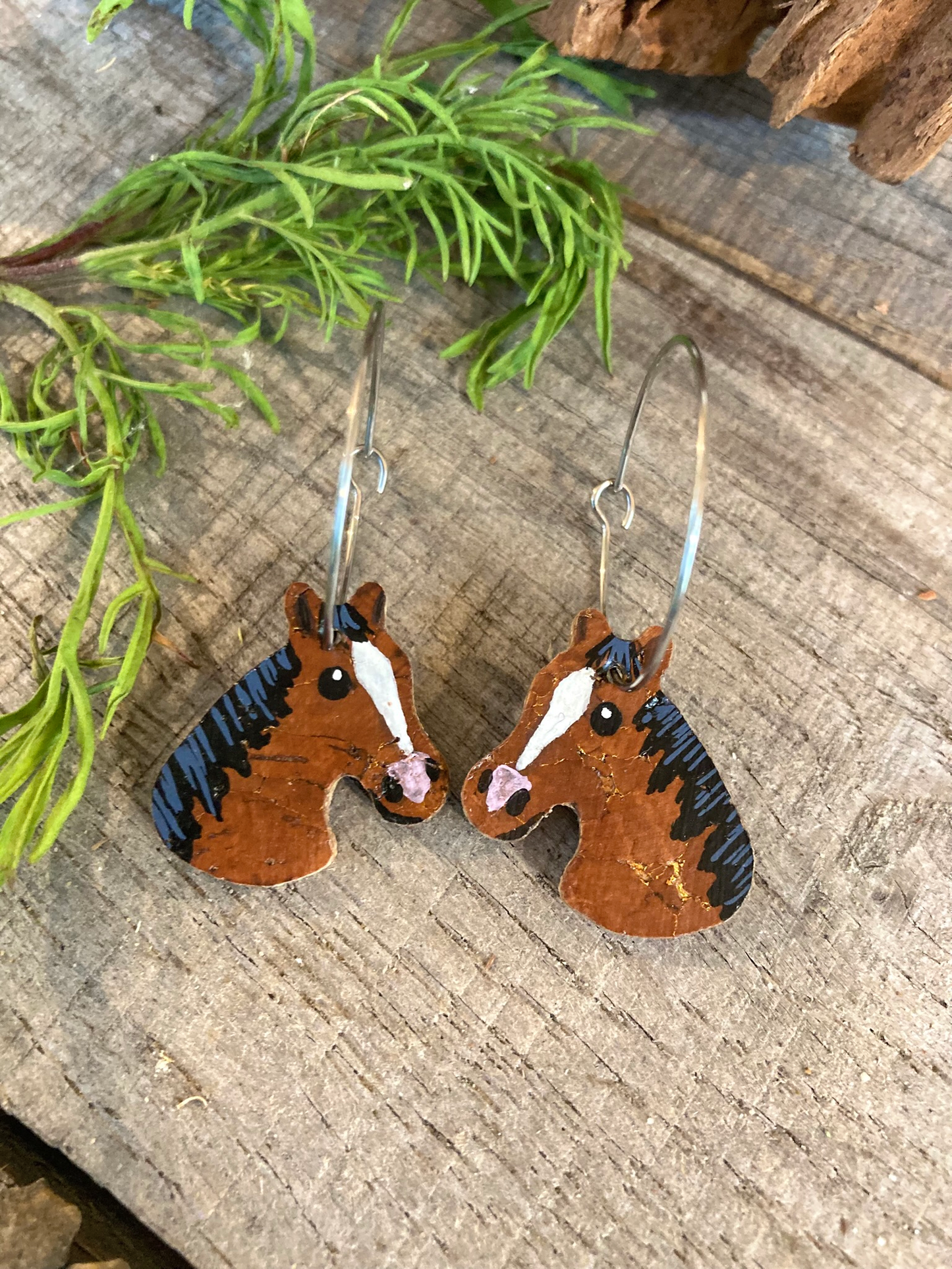 Bay and brown horse cork earrings on silver hoops, flat lay on wooden surface with green foliage — handpainted equestrian jewellery handmade in Suffolk UK