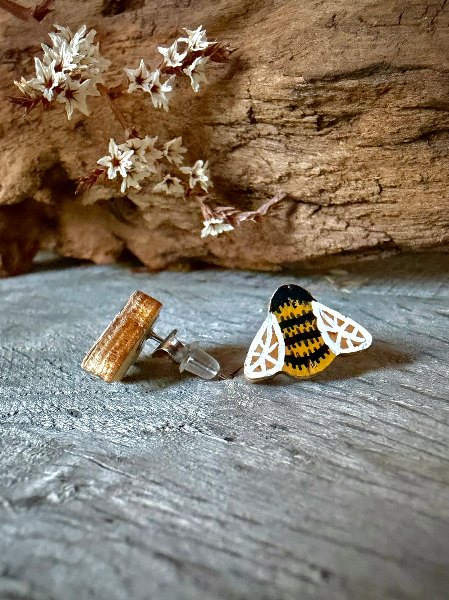 Side view of a bee cork stud earring on weathered wood showing the stainless steel post and silicone back against dried white blossom