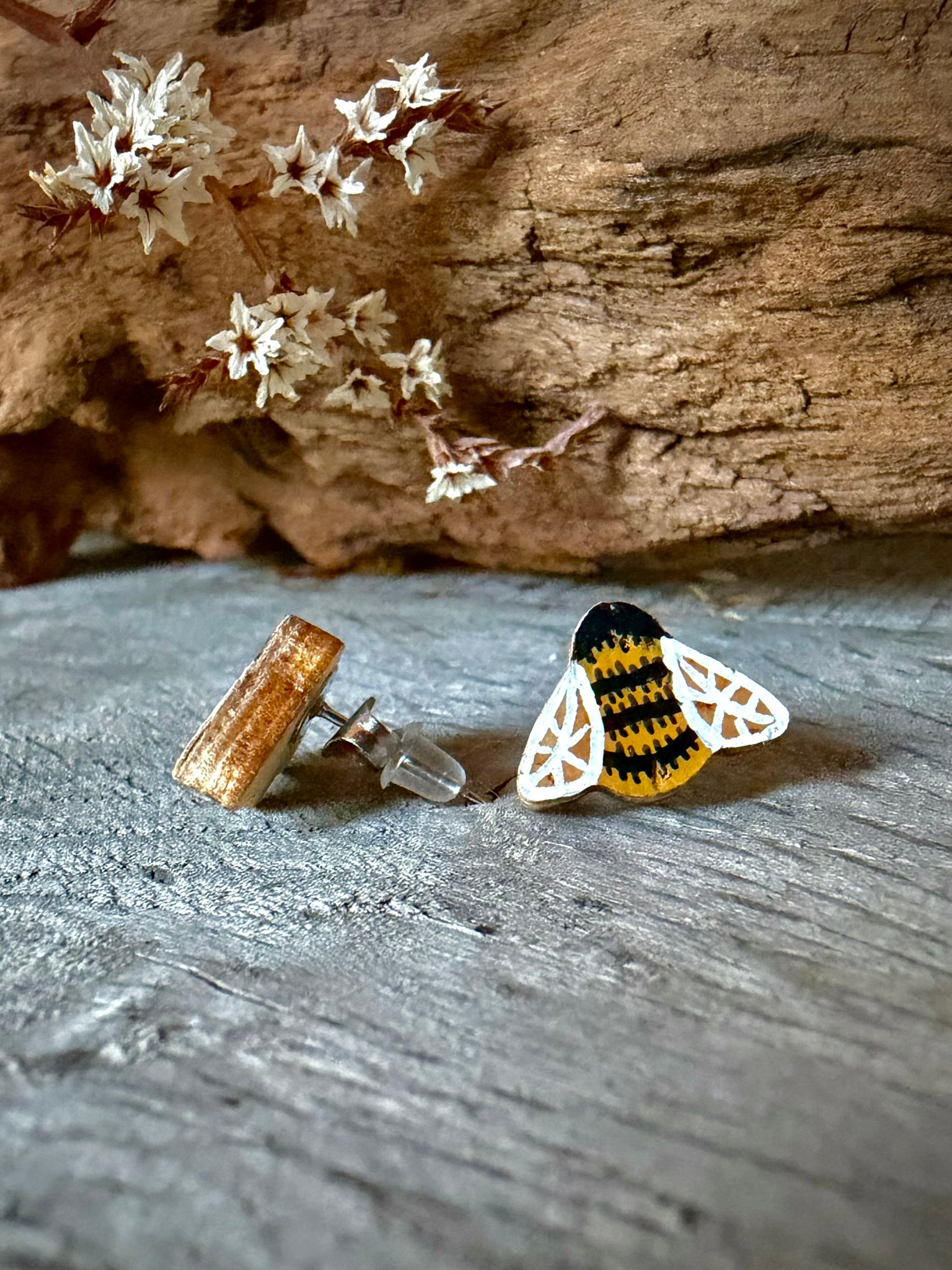 Side view of a bee cork stud earring on weathered wood showing the stainless steel post and silicone back against dried white blossom