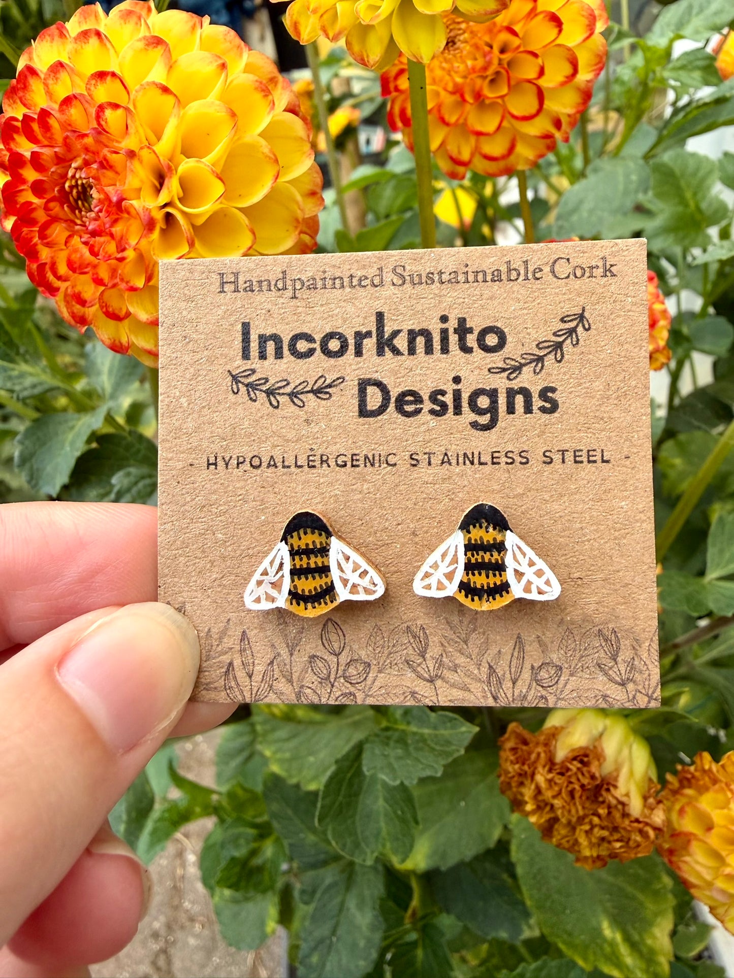 Handpainted bee cork stud earrings on an Incorknito Designs kraft card held against vibrant orange and yellow dahlias