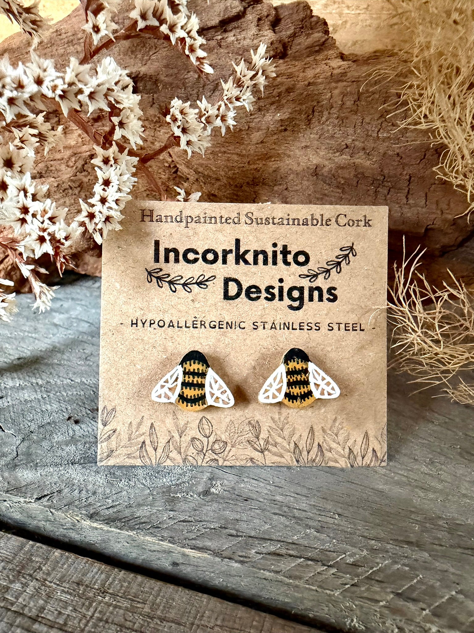 Handpainted bee cork stud earrings on an Incorknito Designs kraft card against driftwood and dried white blossom