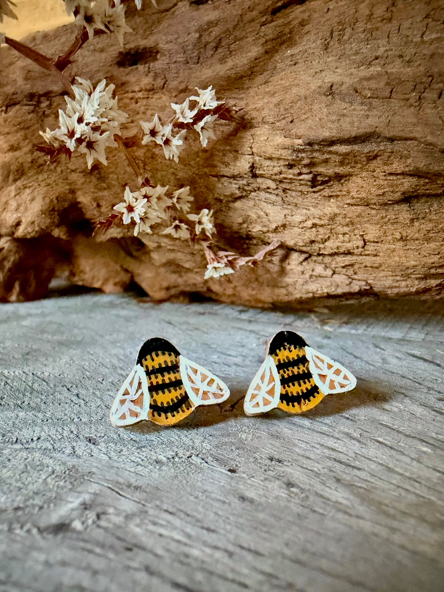Pair of bee cork stud earrings on a weathered wood surface with dried white blossom