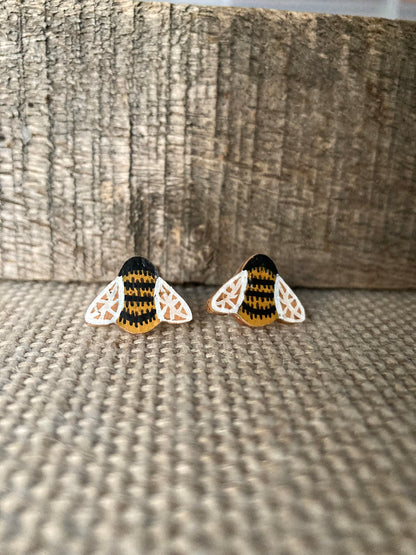 Pair of bee cork stud earrings on a linen surface against weathered wood, showing yellow ochre and black striped body and white filigree wings