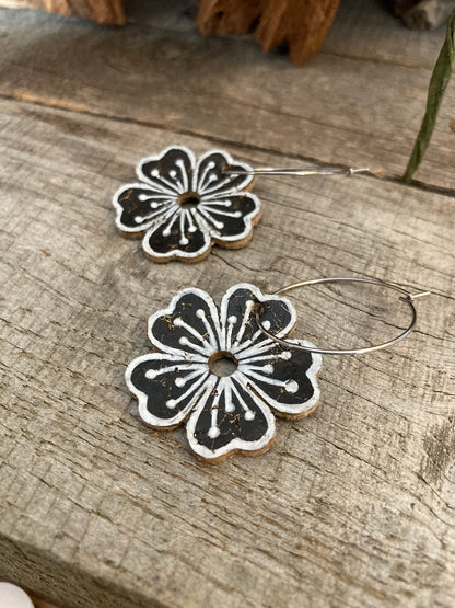 Black large flower cork hoop earrings flat lay on weathered wood with dried botanicals — silver hoops showing gold fleck cork texture and handpainted white pollen detail