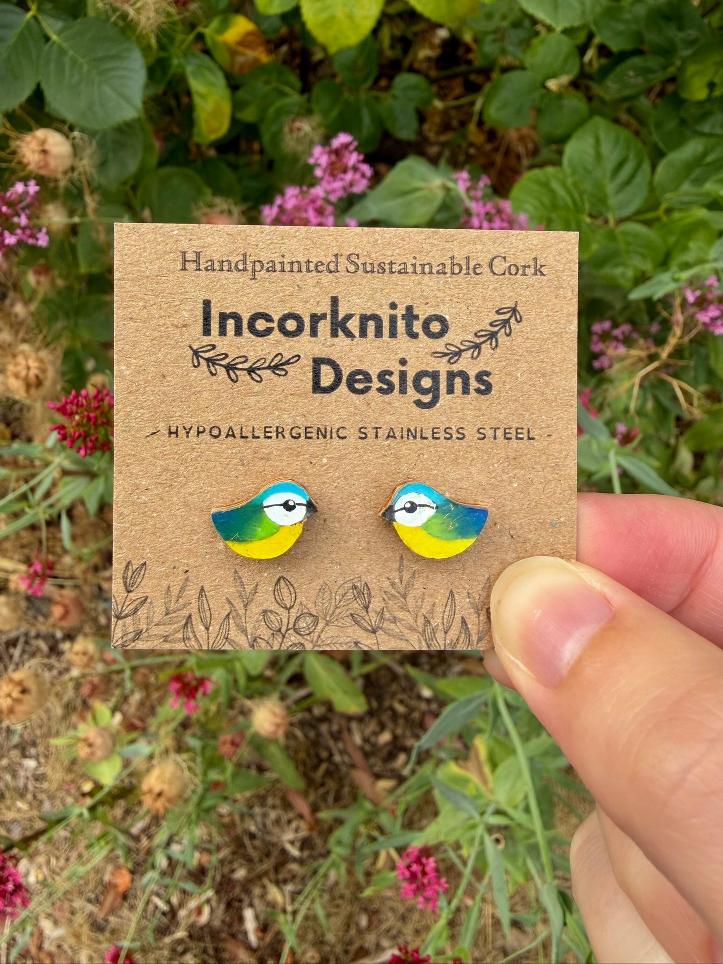 Handpainted blue tit stud earrings held on an Incorknito Designs card against a garden background with pink flowers