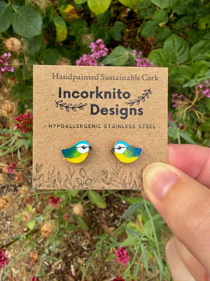 Handpainted blue tit stud earrings held on an Incorknito Designs card against a garden background with pink flowers