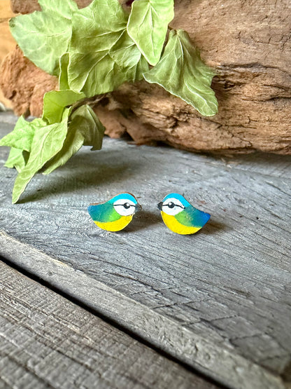 Handpainted blue tit cork stud earrings on a weathered wood surface with ivy, showing sky blue crown, yellow breast and fine black eye detail