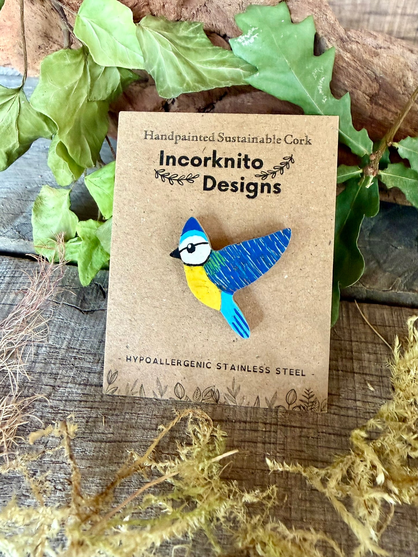 Blue tit cork pin badge on Incorknito Designs branded card against driftwood, ivy and oak leaves — handpainted sustainable cork badge