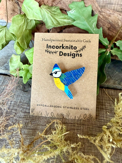 Blue tit cork pin badge on Incorknito Designs branded card against driftwood, ivy and oak leaves — handpainted sustainable cork badge