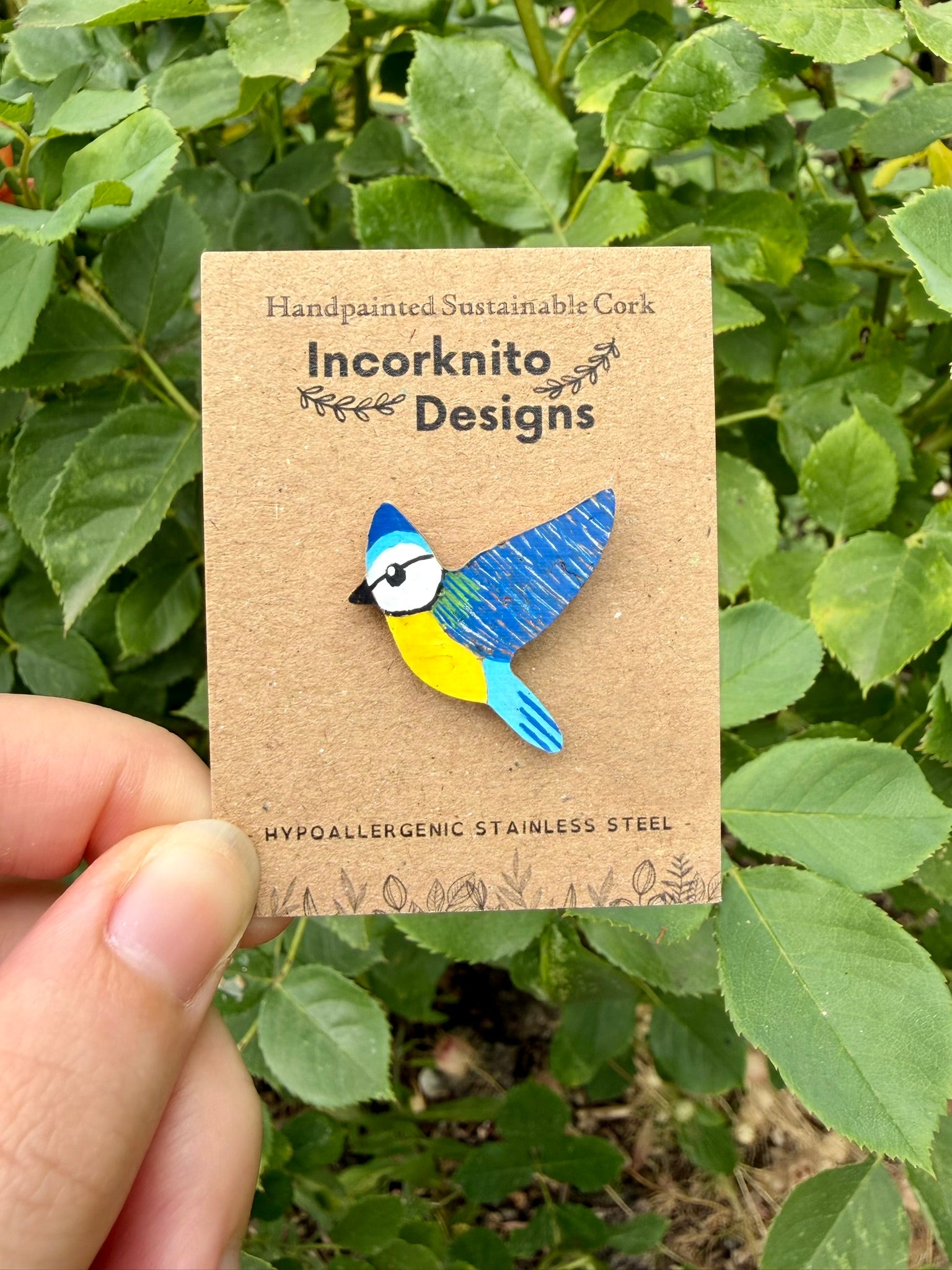 Blue tit cork pin badge on Incorknito Designs branded card held against green garden foliage — handpainted sustainable cork badge handmade in Suffolk UK